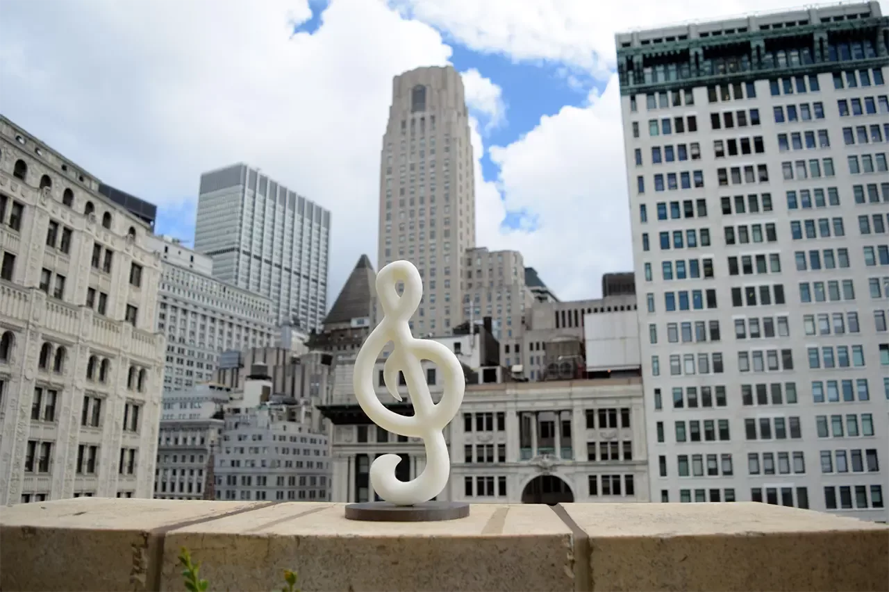 A magnetic 3 dimensional treble clef sculpture sitting on a metal base, with New York City in the background.